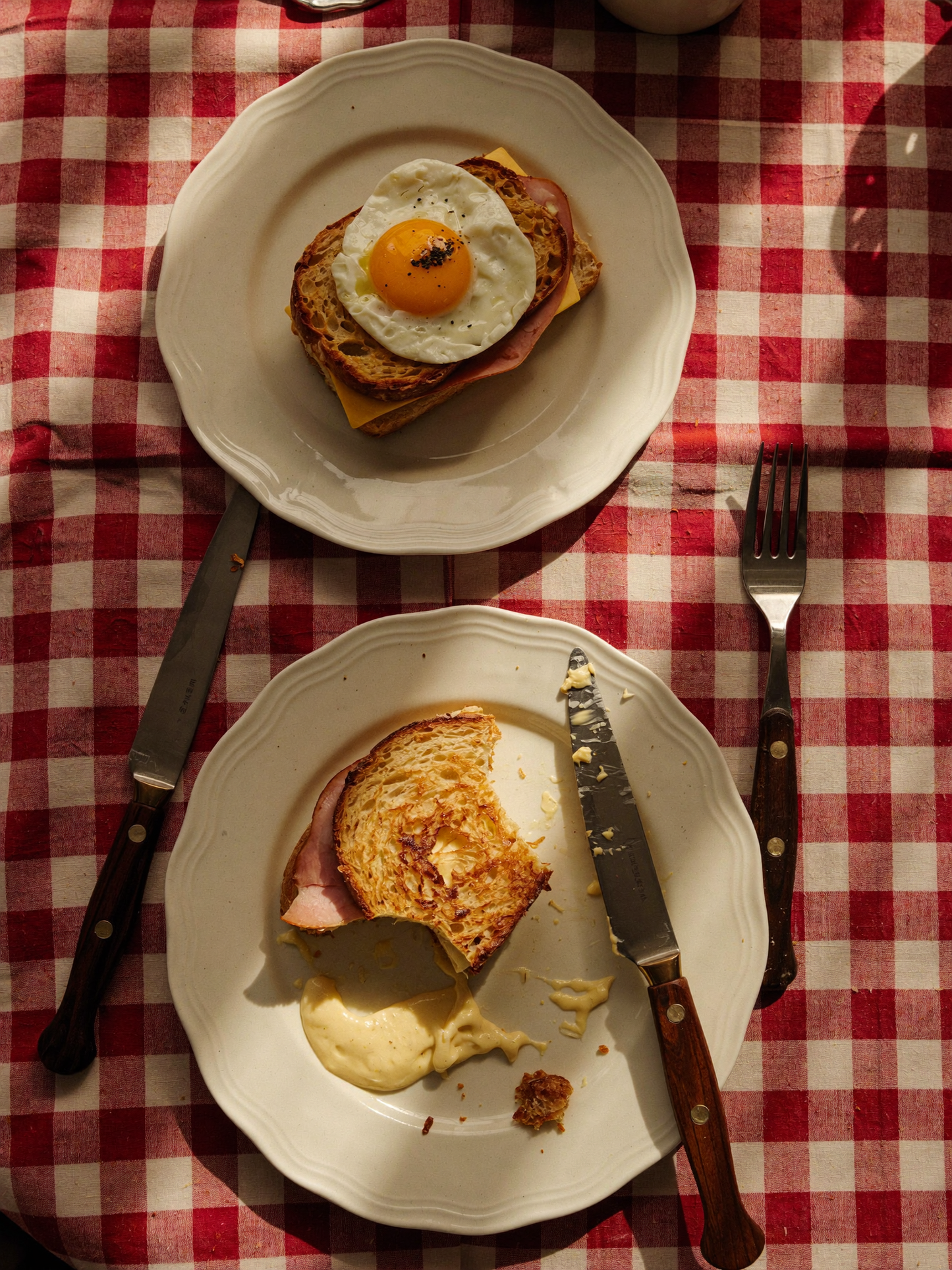 To rustikke morgenmadssmørrebrød på hvide tallerkener, serveret på en rød- og hvidternet dug. Det ene toppet med spejlæg, det andet med smeltet ost og skinke, omgivet af bestik i varmt morgenlys.