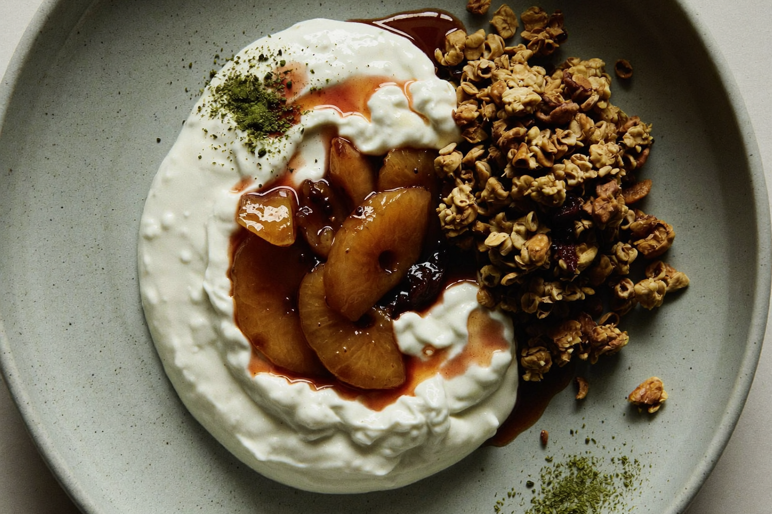 Top-down view of a ceramic bowl with creamy yogurt topped with stewed pear slices and syrup, a cluster of granola on the side, and a light sprinkle of green powder, set on a neutral surface in soft natural light.
