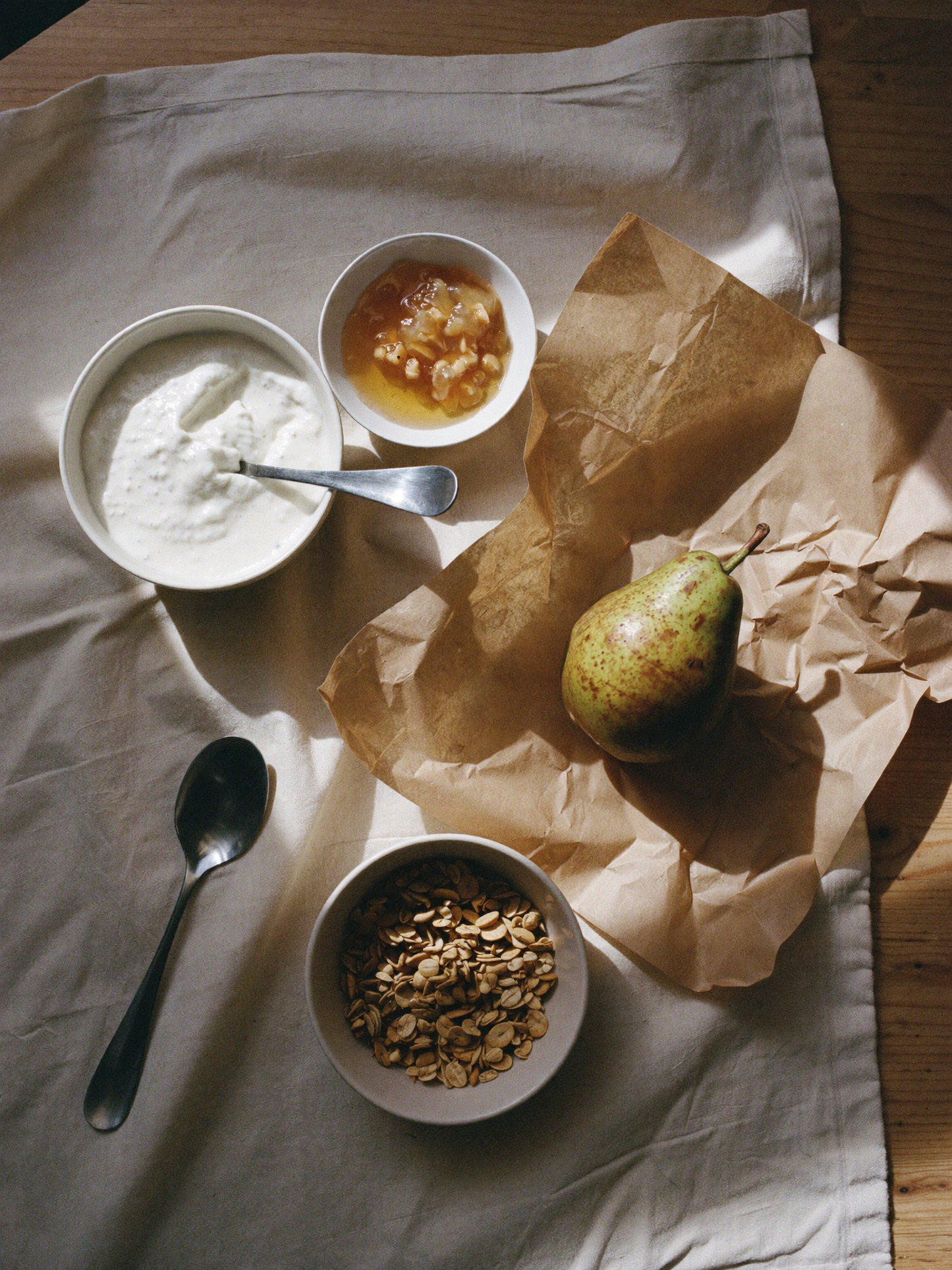 Enkel morgenmadsopstilling med yoghurt, honning, granola og en pære på bagepapir, serveret på en lys dug i blødt, naturligt morgenlys.